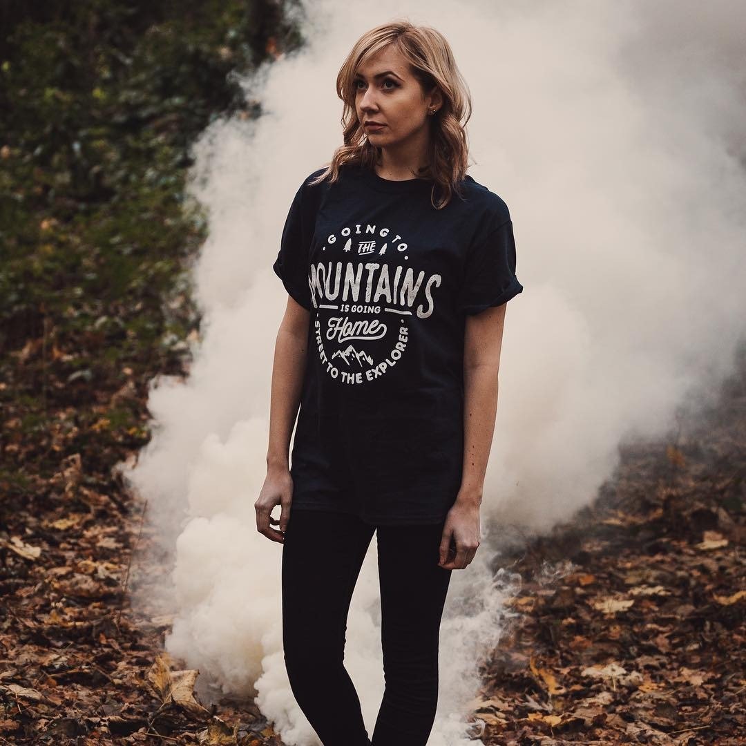 Black organic cotton tee with white text "Going to the Mountains Is Going Home" worn outdoors with smoke and foliage.