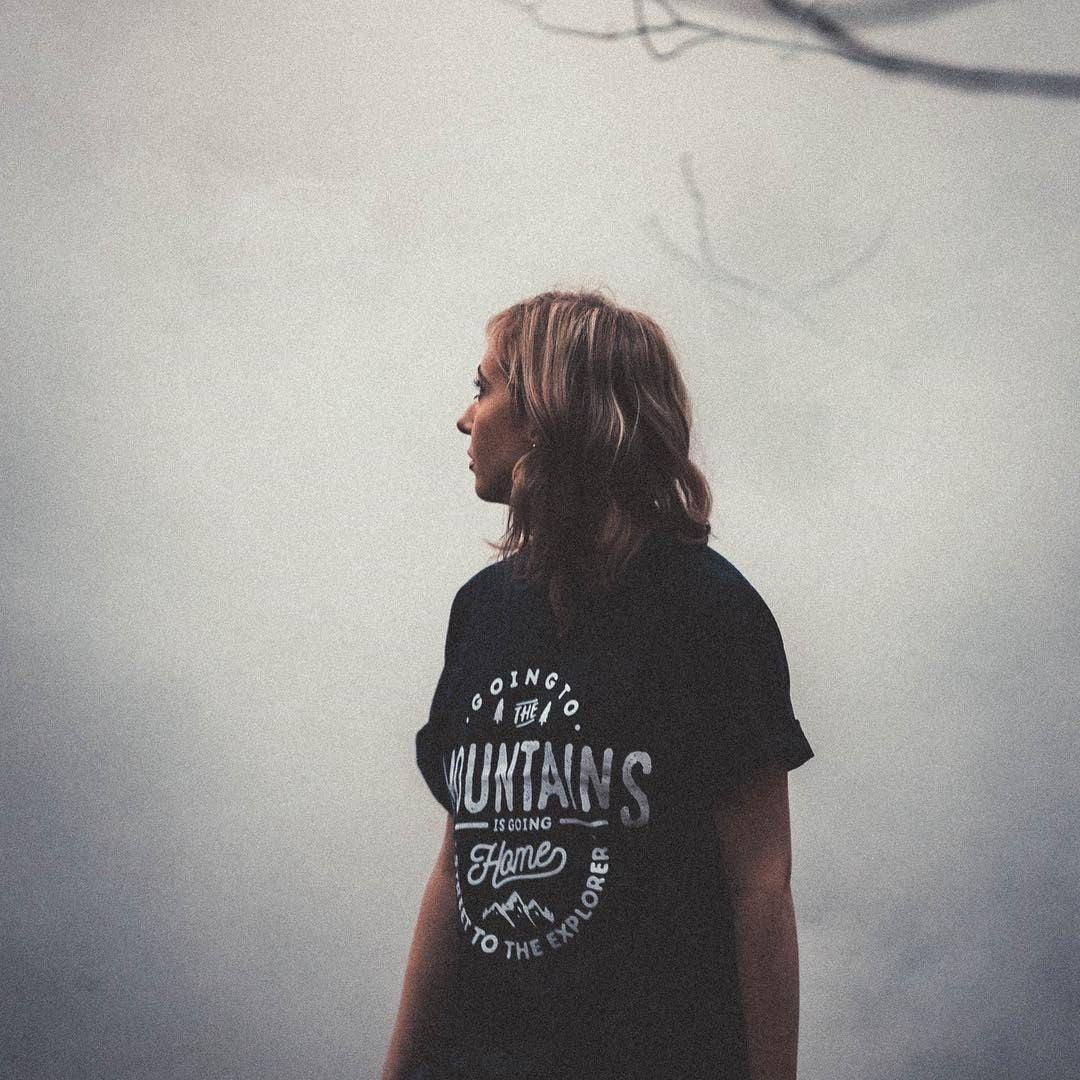 Person wearing a black t-shirt with text and mountain graphic, standing against a foggy background.